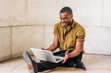 New York 'ta okuyan, yeşil kısa kollu gömlekli, siyah pantolonlu, deri ayakkabılı, mermer zeminde oturan, dizüstü bilgisayarda çalışan, gülümseyen genç bir Afrikalı Amerikalı.