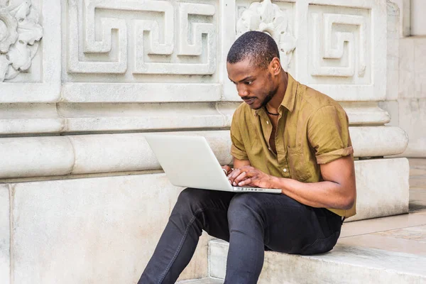 Young African American College Student studying in New York City, wearing green short sleeve shirt, black pants, sitting on stairs outside office building on campus, working on laptop computer