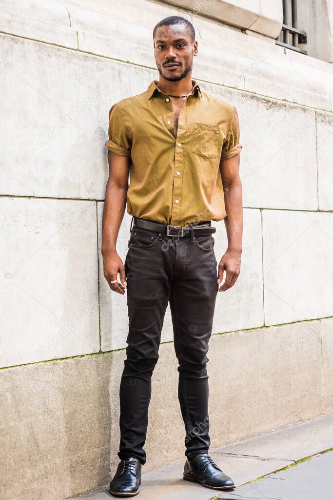 Portrait of Young African American Male College Student in New York City, with beard, wearing green short sleeve shirt, black pants, leather shoes, standing outside office building against wall