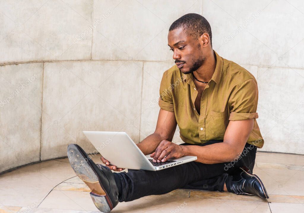 Young African American Man with beard studying in New York, wearing green short sleeve shirt, black pants, leather shoes, sitting legs crossed on marble ground on campus, working on laptop computer