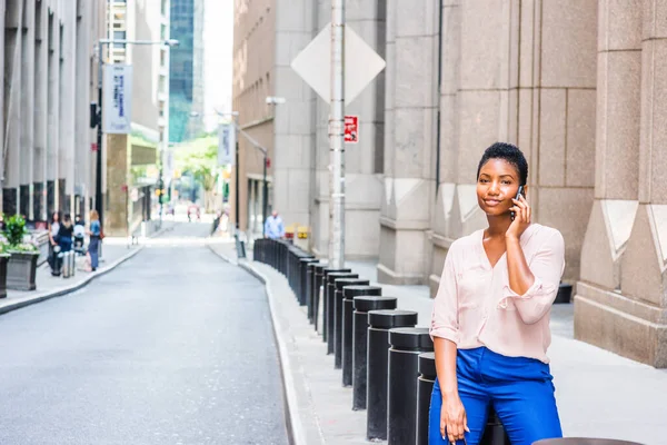 Young African American Woman with short afro hair, traveling in New York City, wearing pink v neck shirt, blue pants, sitting on black metal pillar on narrow street, talking on cell phone, smiling