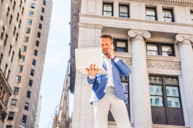 Sakallı genç Avrupalı iş adamı New York 'ta çalışıyor, mavi ceket giyiyor, gri pantolon giyiyor, eski ofis binasının önünde duruyor, dizüstü bilgisayarla çalışıyor, cep telefonuyla konuşuyor.