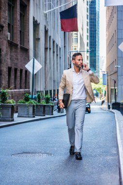 Avrupalı iş adamı seyahat ediyor, New York 'ta çalışıyor, sakallı, kır saçlı, bej ceketli, gri pantolonlu, deri ayakkabılı, evrak çantalı, sokakta yürüyor, cep telefonuyla konuşuyor.