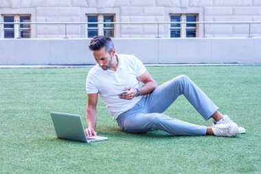 Sakallı, kısa kollu beyaz gömlekli, gri pantolonlu, beyaz spor ayakkabılı, New York 'ta yeşil çimlerde oturan, dizüstü bilgisayarda çalışan, cep telefonuyla mesajlaşan genç bir Avrupalı.