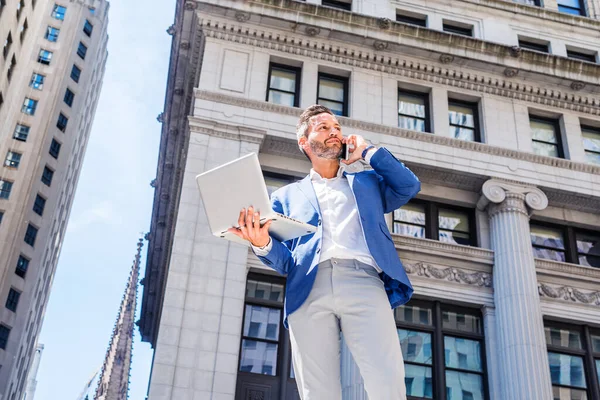 Sakallı genç Avrupalı iş adamı New York 'ta çalışıyor, mavi ceket giyiyor, gri pantolon giyiyor, eski ofis binasının önünde duruyor, dizüstü bilgisayarla çalışıyor, cep telefonuyla konuşuyor.