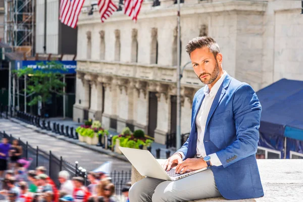 Young European Businessman with beard traveling, working in New York City, wearing blue blazer, sitting on street outside office building, working on laptop computer, looking up, thinking