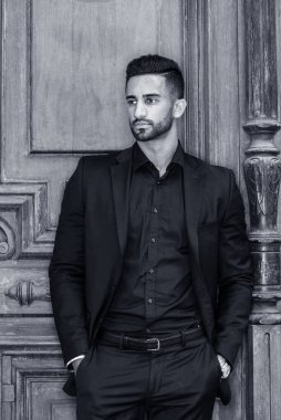 New York 'ta sakallı, siyah takım elbiseli, elleri ceplerinde, eski tarz ofis kapısının önünde dikilen genç, Doğu Hintli Amerikalı iş adamının portresi. Siyah ve Beyaz