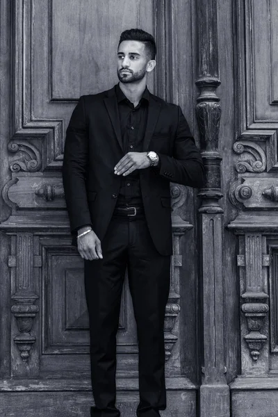 Young East Indian American Businessman with beard, wearing black suit, wristwatch, standing by old style office doorway in New York City, hand in front touching clothes, looking away. Black and White