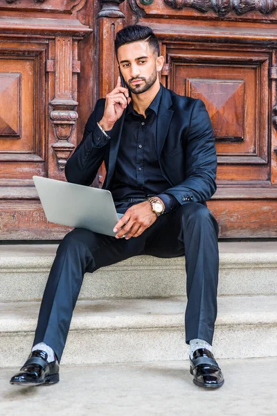 New York 'ta sakallı, siyah takım elbiseli, siyah gömlekli, kol saati takan, ofis kapısının önünde oturan, dizüstü bilgisayarla çalışan, cep telefonuyla konuşan Doğu Hintli genç bir Amerikalı.