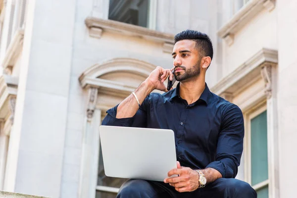 Young East Indian American Businessman with beard working in New York City, wearing black shirt, holding laptop computer, sitting outside old style office building on campus, talking on cell phone