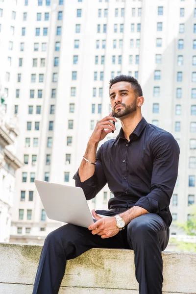 Young East Indian American Businessman with beard traveling, working in New York City, wearing black shirt, holding laptop computer, sitting outside old style office building on street, talking on cell phone