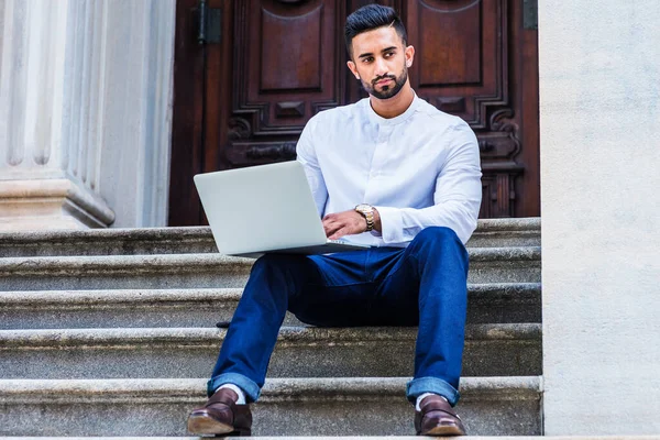 New York 'ta sakallı, beyaz gömlekli, mavi pantolonlu, siyah deri ayakkabılı, ofisin dışındaki merdivenlerde oturan, dizüstü bilgisayarla çalışan genç Doğu Hint Amerikan Koleji öğrencisi.