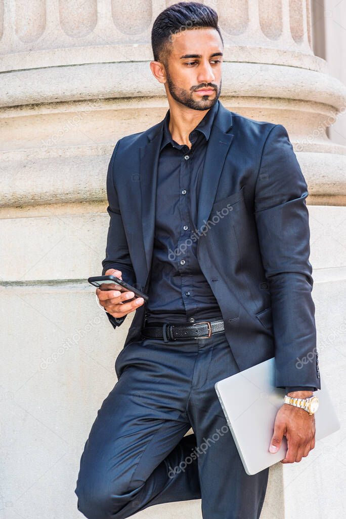 Young East Indian American Businessman with beard working in New York, wearing black suit, carrying laptop computer, standing by column outside office, holding cell phone, looking away,  thinking