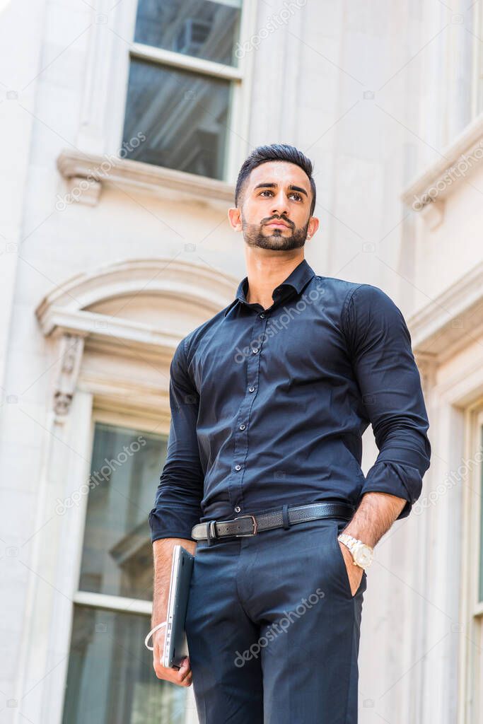 Portrait of Young East Indian American Businessman with beard in New York City, wearing black suit, black pants, carrying laptop computer, standing outside old style office building, looking forward