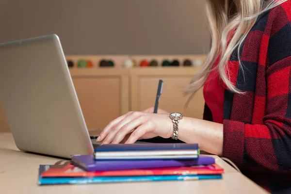 Blonde woman sitting at a laptop and writing. There is a laptop, a tablet, a phone and a notebook on the table.