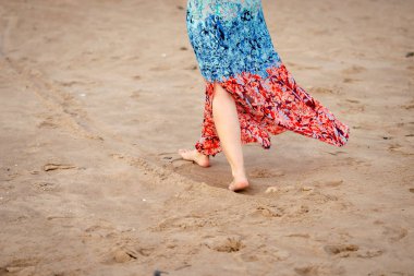 Çıplak ayak ve plaj boyunca yürüyüş renkli uzun elbisesi olan bir genç kadın. Sadece gövdenin alt kısmında görünür.