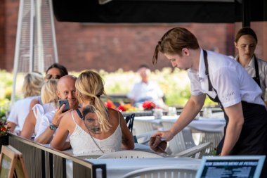 Riga, Letonya - 31 Temmuz 2018: Masada açık kafe sırtında büyük bir dövme olan bir kadın oturuyor. Garson ziyaretçi hizmet vermektedir..