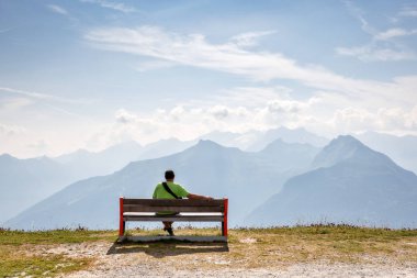 Bir adam Alpler üst kısmında ahşap bir bankta oturur ve güzel dağ manzaraya görünüyor.