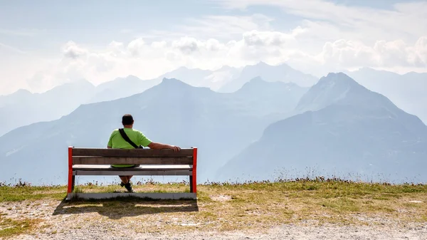 A man sits on a wooden bench at the top of the Alps and looks at the beautiful mountain landscape.