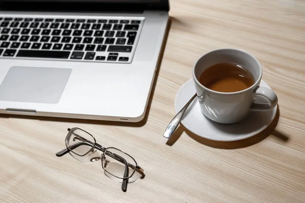 Desk with laptop, eyeglasses and a cup of tea on a wooden table  - image
