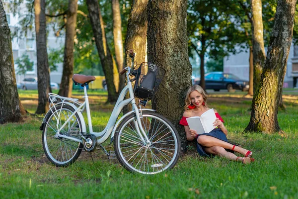 A young blonde woman with a white bike is sitting in a park near a tree and reading a book - image