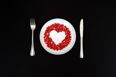 Nar tohumları bileşik kalp şekli beyaz bir plaka üzerinde. Bıçak ve çatal plaka yanında. Siyah arka plan. Düz tabaka. 