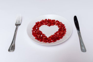 Nar tohumları bileşik kalp şekli beyaz bir plaka üzerinde. Bıçak ve çatal plaka yanında. Beyaz arka plan.