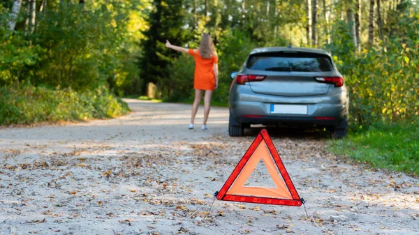 Stressful young woman driver hitchhikes and stops cars, asks for help as have problem with brocken car, uses red triangle sign to warn drivers about stop.  (selective focus)