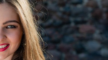 Güneşli bir günde eski bir taş duvarın yanında gülümseyen genç bir kadın.