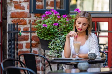 Açık renk elbiseli güzel sarışın kadın açık hava kafede oturup kokteyl yudumluyor.. 