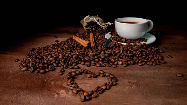 White coffee mug, cinnamon sticks  and coffee beans on the dark wooden background, Heart shape from coffee beans in the foreground - image