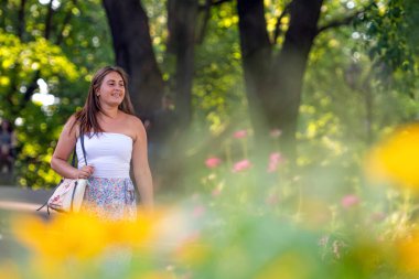 Açık bluz giyen ve değişik etek giyen genç bir kadın şehir parkında yürüyor, ön plandaki çiçekleri dağıtıyor.