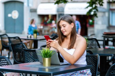 Genç kadın dizüstü bilgisayarda çalışıyor ve telefon kullanıyor sokak kafesinde oturuyor, uzaktan öğrenme ve çalışma konsepti