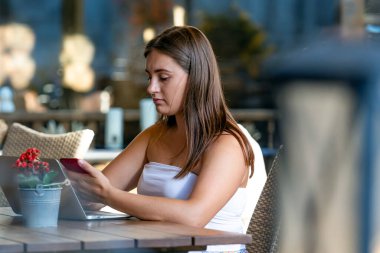 Genç kadın dizüstü bilgisayarda çalışıyor ve telefon kullanıyor sokak kafesinde oturuyor, uzaktan öğrenme ve çalışma konsepti