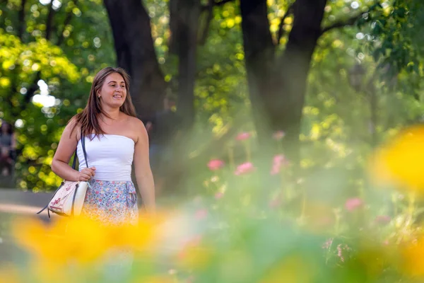 Açık bluz giyen ve değişik etek giyen genç bir kadın şehir parkında yürüyor, ön plandaki çiçekleri dağıtıyor.