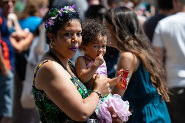 New York, ABD - 22 Haziran 2019: Kafasında çiçek çelengi ve ellerinde bir bebek olan Afrikalı kadın sokakta kalabalıkta yürüyor