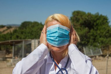 Sarışın kadın doktor göz ve kulak cerrahisi maskesi takıyor. Virüs hakkında olumsuz şeyler duymak ya da görmek istememenin sembolü olarak.