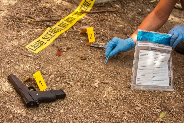 close up of a detective hand removing evidence of the scene like gun or bloody knife from the crime scene numbered with yellow cards and yellow police tape and placing the evidence in the appropriate bag