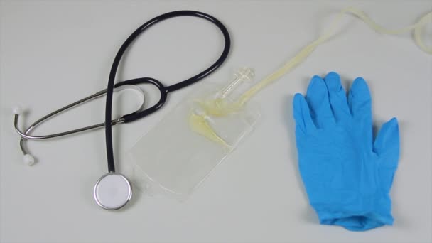 Katéter vízelvezető zsák, amely megtelik vizelettel elhelyezett kórházi asztalra egy kék kesztyű és sztetoszkóppal mellette — Stock videók