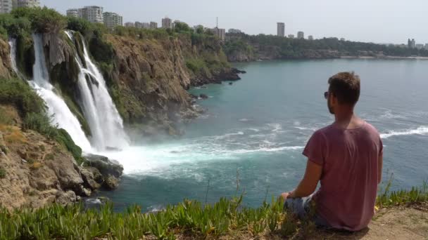 Selfie man. Uomo seduto sulla scogliera vicino alla cascata e gode della vista — Video Stock