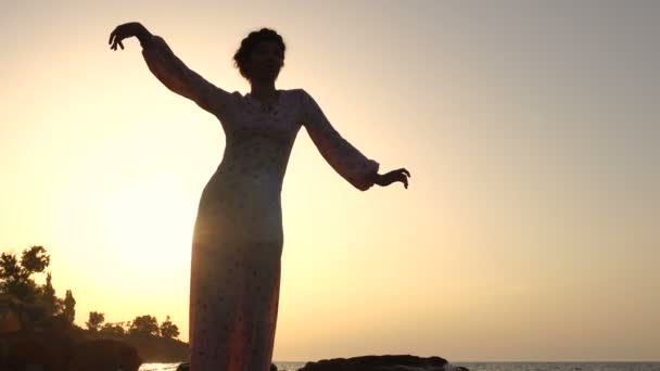 Schattig Kaukasische Brunette meisje in licht jurk dansen op de klif boven de zee bij zonsopgang — Stockvideo