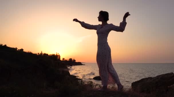 Очаровательная кавказская брюнетка в светлом платье танцует на скале над морем на восходе солнца — стоковое видео