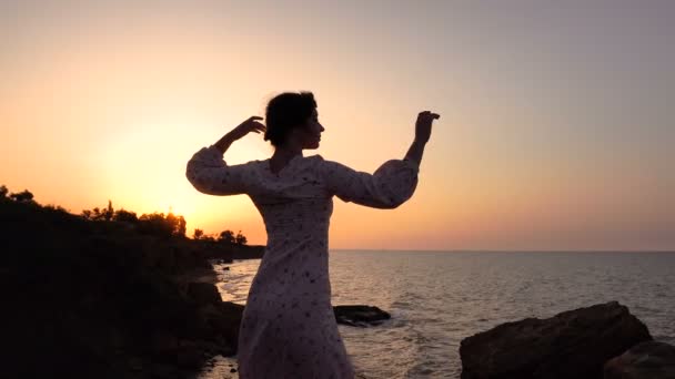 Entzückende kaukasische Brünette in leichtem Kleid, die bei Sonnenaufgang an der Klippe über dem Meer tanzt — Stockvideo