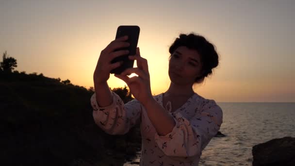 Adorabile ragazza bruna caucasica in abito leggero fai selfie sulla scogliera sopra il mare all'alba — Video Stock