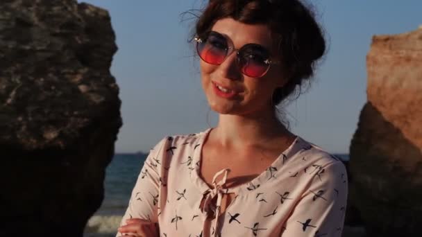 Entzückende kaukasische Brünette mit Sonnenbrille und hellrosa Kleid sitzt bei Sonnenaufgang auf dem Stein — Stockvideo