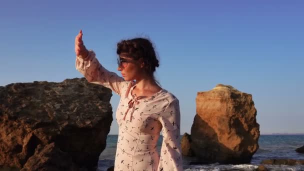 Adorable chica morena caucásica en gafas de sol y vestido rosa claro sentarse en la piedra al amanecer — Vídeos de Stock