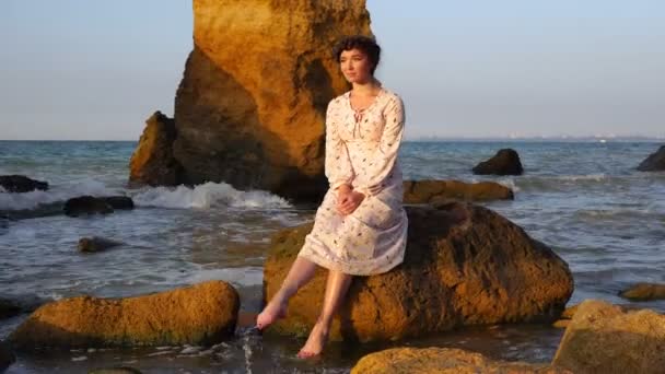 Schattig Kaukasische Brunette meisje in licht roze jurk zitten op de rots in de zee bij zonsopgang — Stockvideo