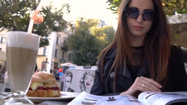 Aantrekkelijke moderne jonge vrouw drinkt aromatische koffie in een café op een zomerterras en een blad over mode-tijdschrift — Stockvideo