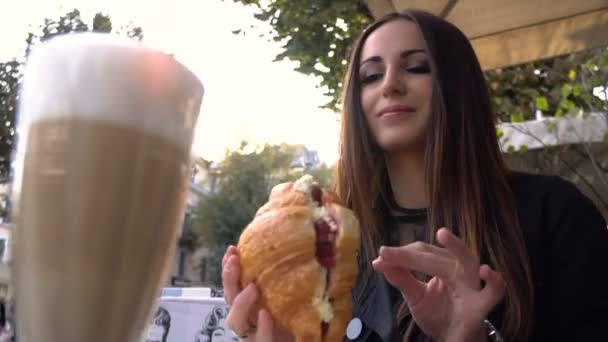 Aantrekkelijke moderne jonge vrouw croissantje eten en drankjes heerlijk geurende koffie in een café op een zomerterras. — Stockvideo