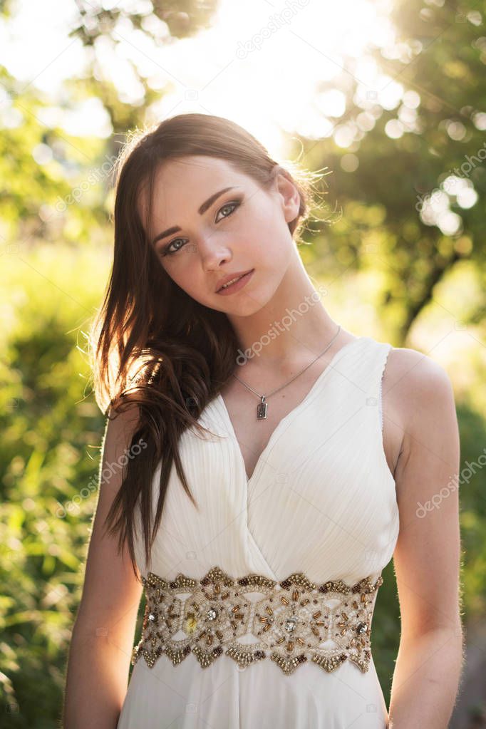 Summer portrait of young beautiful lady wearing long white evening dress posing in the park.